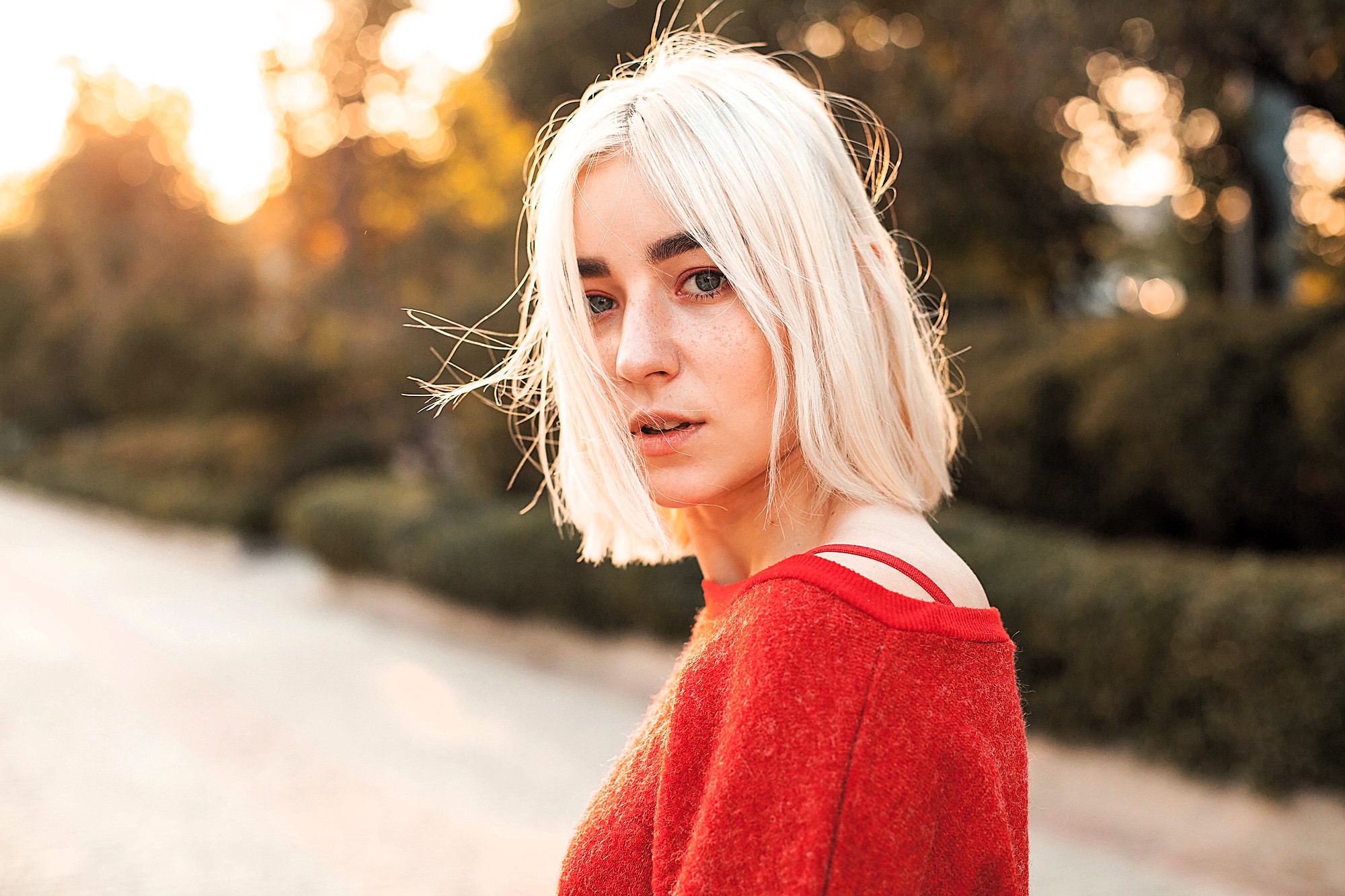 A young person with short, blonde hair stands outdoors. They are wearing a red sweater and looking at the camera. The background is softly blurred, with warm sunlight filtering through trees.