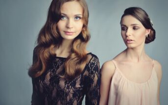 Two women stand against a gray background. The woman on the left has long wavy hair and wears a black lace top. The woman on the right has her hair in an updo and wears a light pink dress, looking towards the other woman.