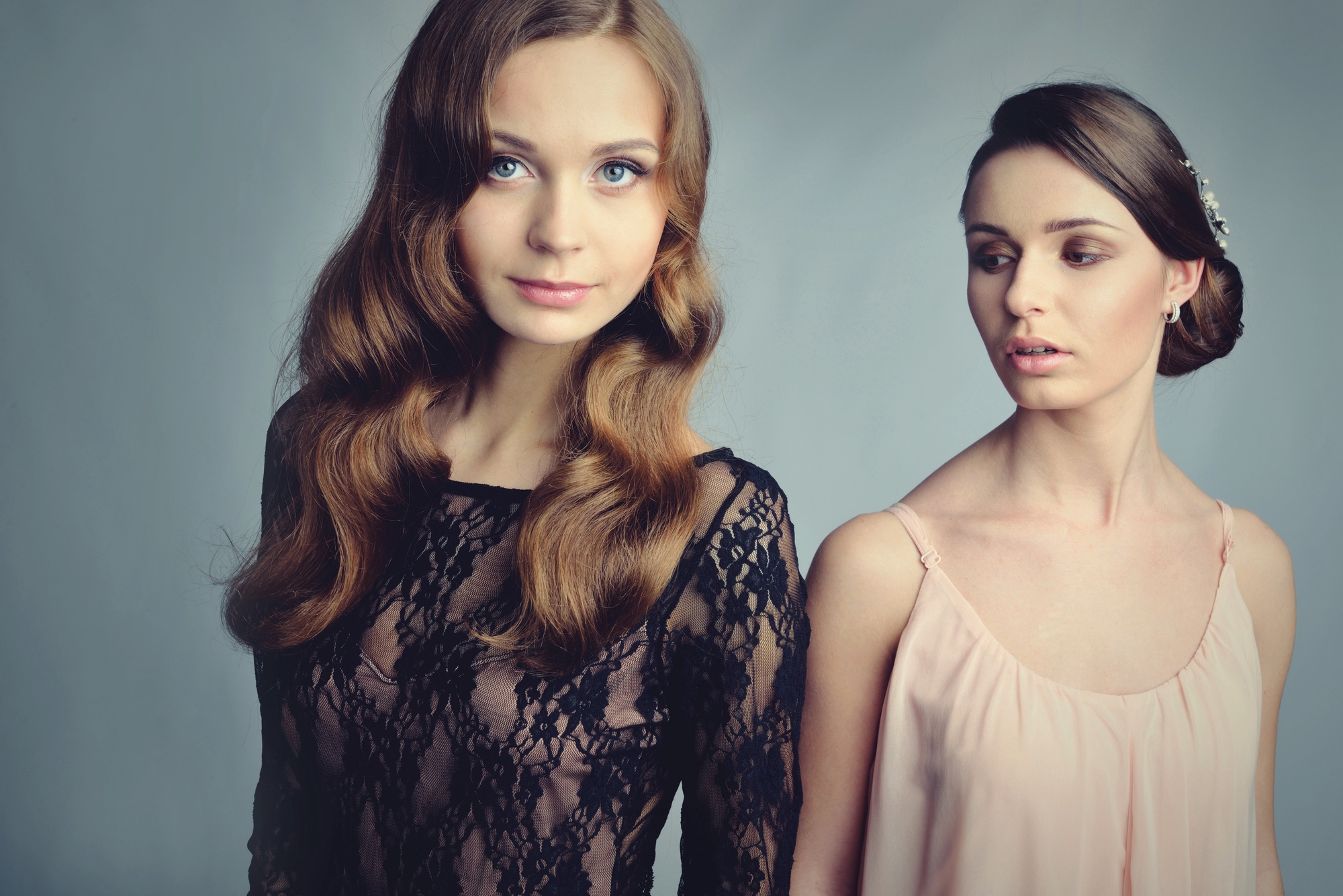 Two women stand against a gray background. The woman on the left has long wavy hair and wears a black lace top. The woman on the right has her hair in an updo and wears a light pink dress, looking towards the other woman.