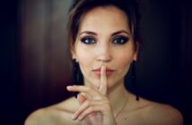 A woman with light skin and dark hair holds her finger to her lips in a shushing gesture. Her expression is calm, and she wears dark eye makeup and dangly earrings. The background is softly blurred.