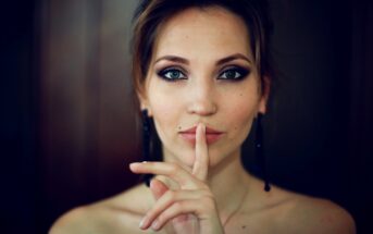 A woman with light skin and dark hair holds her finger to her lips in a shushing gesture. Her expression is calm, and she wears dark eye makeup and dangly earrings. The background is softly blurred.