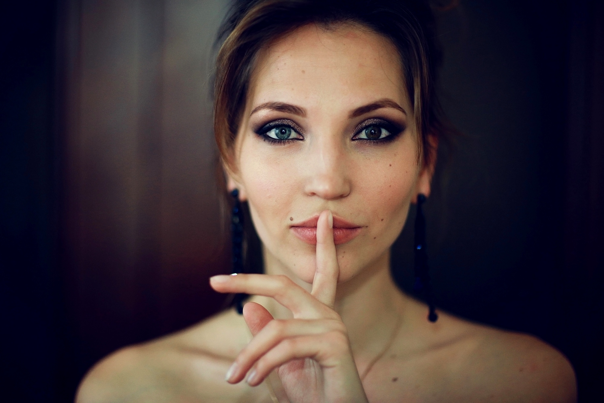 A woman with light skin and dark hair holds her finger to her lips in a shushing gesture. Her expression is calm, and she wears dark eye makeup and dangly earrings. The background is softly blurred.