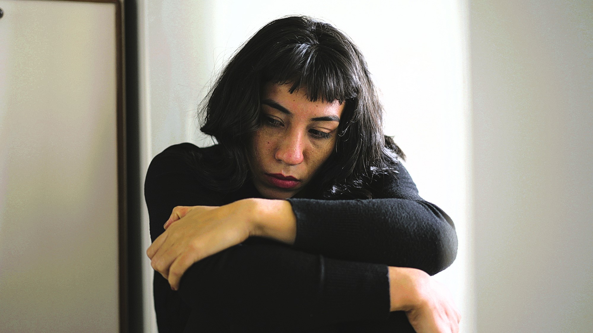 A person with long, dark hair sits pensively with their arms wrapped around their knees, wearing a black top. They are gazing downward, showing a thoughtful or contemplative expression. The background is softly lit, creating a calm atmosphere.