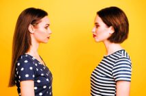 Two women facing each other against a bright yellow background. The woman on the left has long, straight hair and wears a polka dot top, while the woman on the right has a short bob haircut and wears a striped top.