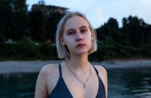 A young woman with short blonde hair gazes at the camera, wearing a dark swimsuit and a necklace. She stands by a calm body of water, with a blurred background of trees and a building at dusk.