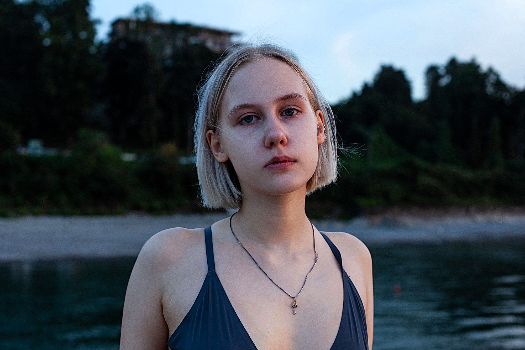 A young woman with short blonde hair gazes at the camera, wearing a dark swimsuit and a necklace. She stands by a calm body of water, with a blurred background of trees and a building at dusk.