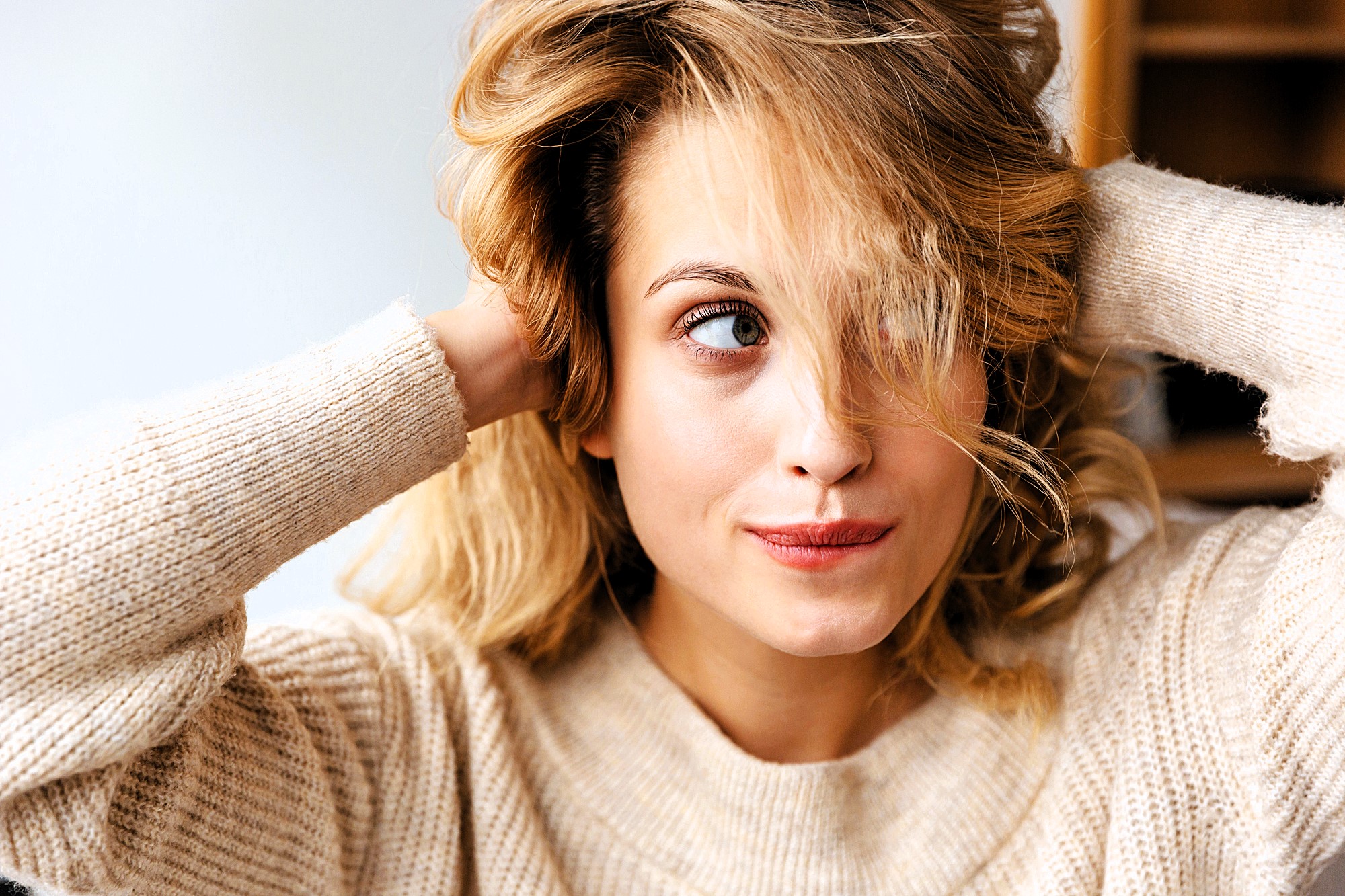 A woman with blonde hair and a slight smile looks off to the side, holding her hands in her hair. She is wearing a beige sweater and is in a softly lit environment.