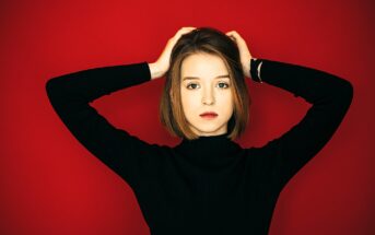 A person with short hair, wearing a black turtleneck, stands against a bright red background with hands on their head, looking directly at the camera.