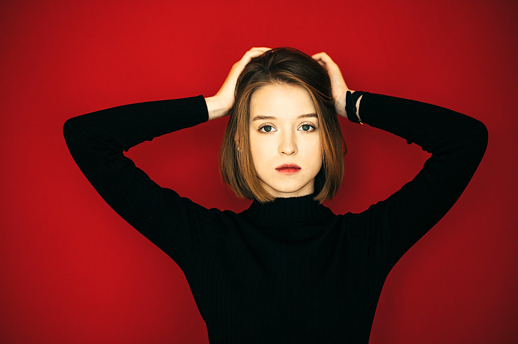 A person with short hair, wearing a black turtleneck, stands against a bright red background with hands on their head, looking directly at the camera.