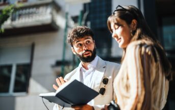 A man with a beard, holding a notebook, gestures while speaking to a woman with long hair. They are outdoors in a sunlit urban setting, with buildings in the background. The woman holds sunglasses and a camera.