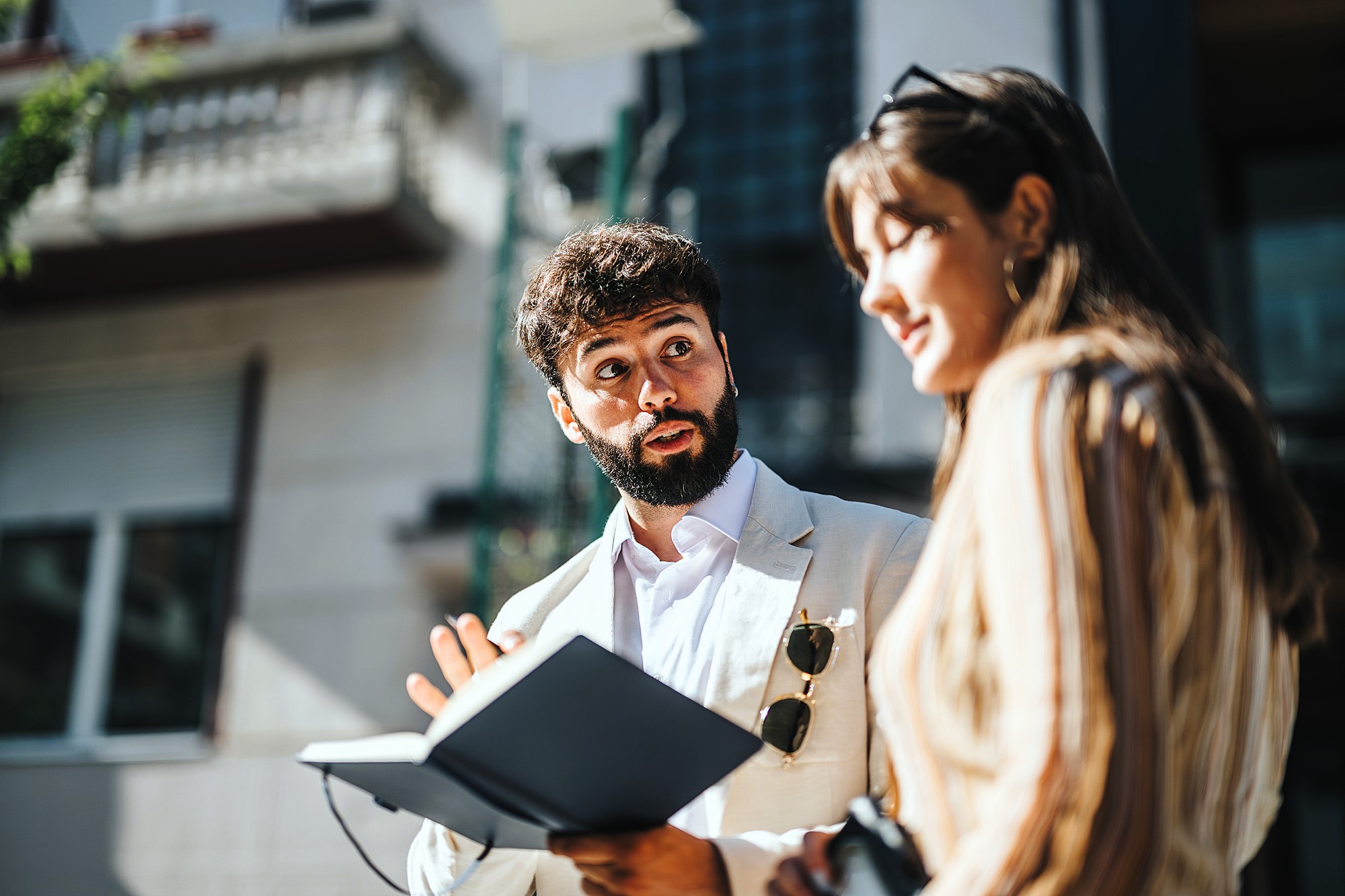 A man with a beard, holding a notebook, gestures while speaking to a woman with long hair. They are outdoors in a sunlit urban setting, with buildings in the background. The woman holds sunglasses and a camera.