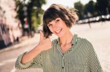 A smiling woman with a bob haircut wearing a green and white striped shirt gives a thumbs-up gesture outdoors on a sunny day. The background shows blurred trees and a street.