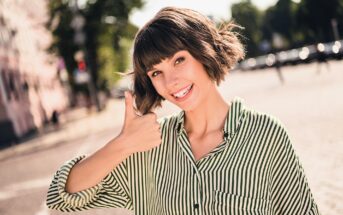 A smiling woman with a bob haircut wearing a green and white striped shirt gives a thumbs-up gesture outdoors on a sunny day. The background shows blurred trees and a street.