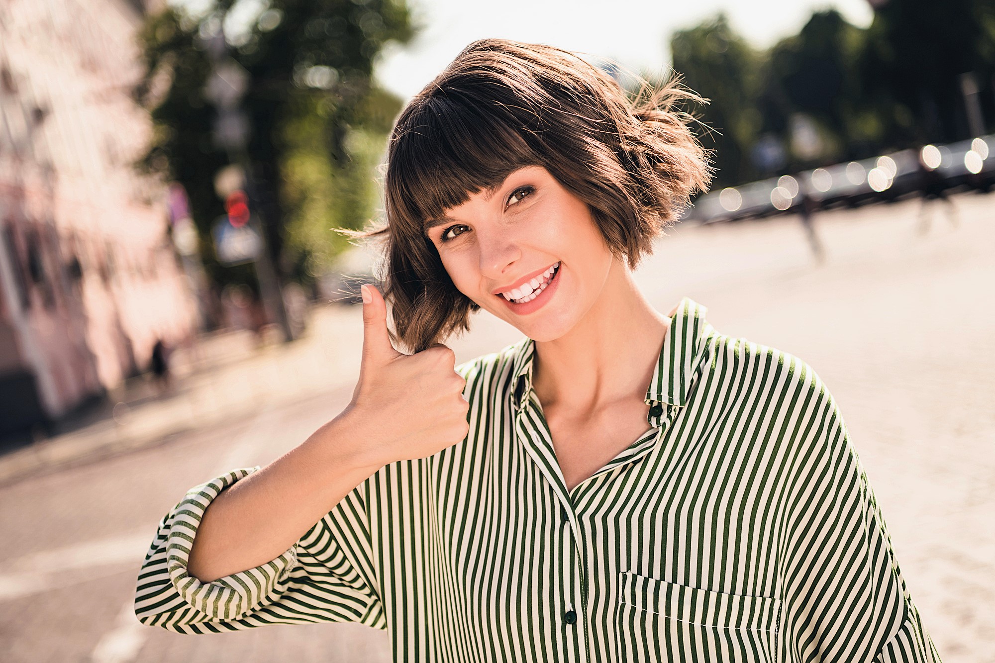 A smiling woman with a bob haircut wearing a green and white striped shirt gives a thumbs-up gesture outdoors on a sunny day. The background shows blurred trees and a street.