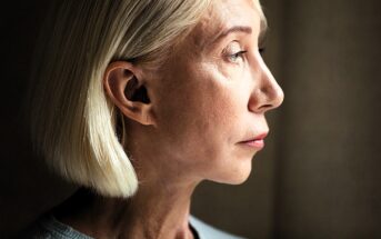 Profile of a person with short, blond hair, looking contemplatively to the side. The lighting highlights the facial features against a dark background.