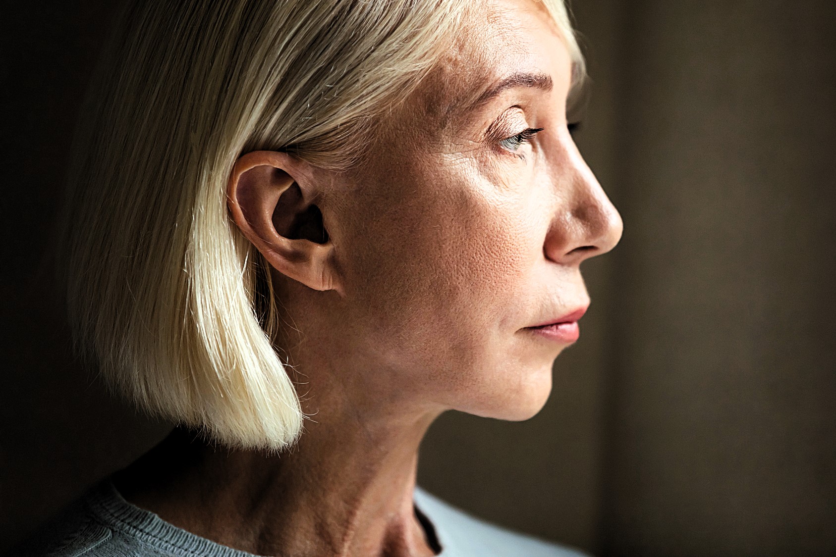 Profile of a person with short, blond hair, looking contemplatively to the side. The lighting highlights the facial features against a dark background.