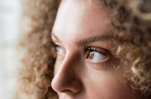 Close-up of a person with curly hair looking to the side. The focus is on the eye and eyebrow, capturing detailed skin texture and eyelashes, with soft natural lighting.