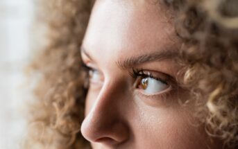 Close-up of a person with curly hair looking to the side. The focus is on the eye and eyebrow, capturing detailed skin texture and eyelashes, with soft natural lighting.