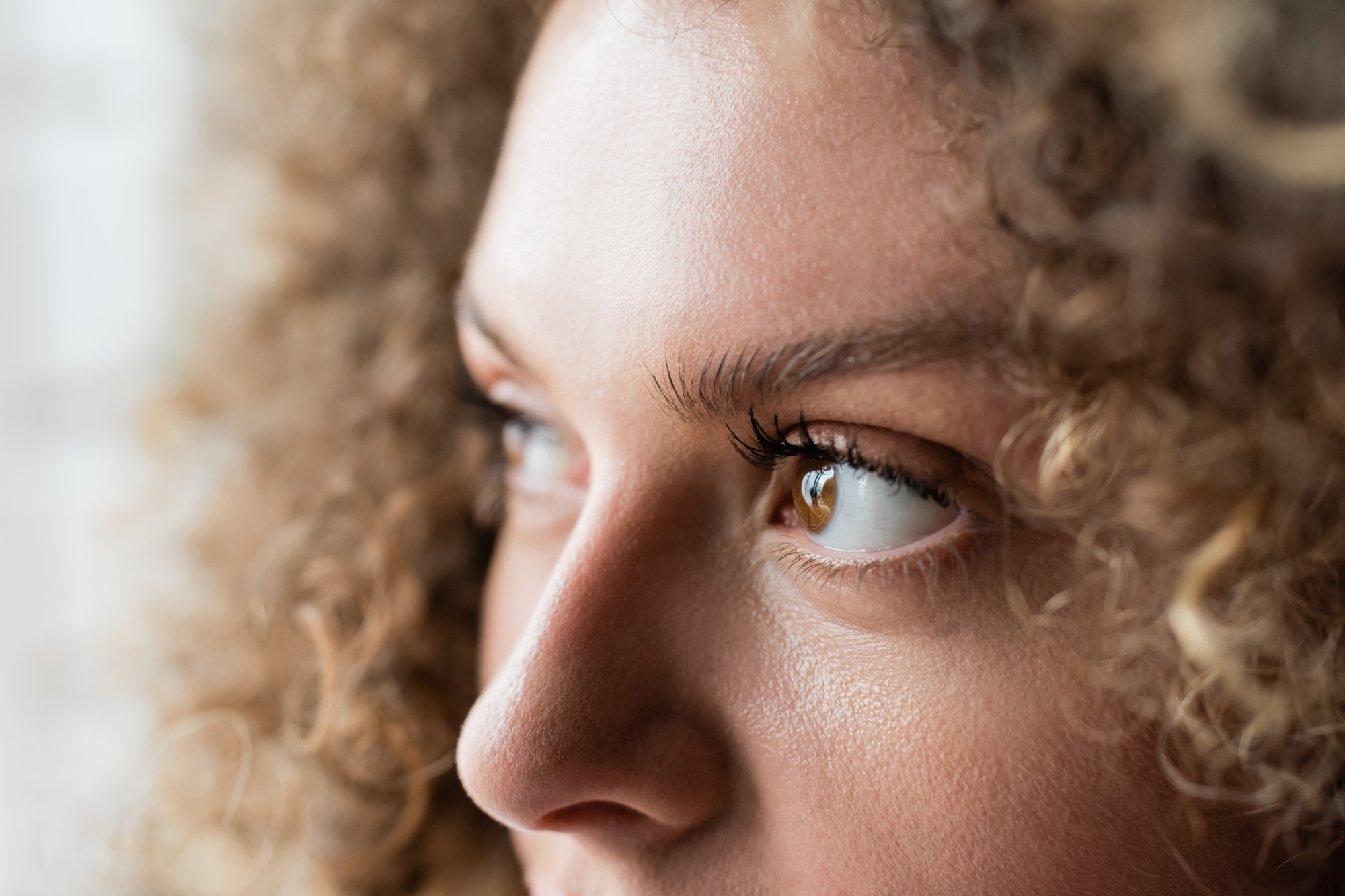 Close-up of a person with curly hair looking to the side. The focus is on the eye and eyebrow, capturing detailed skin texture and eyelashes, with soft natural lighting.