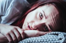 A woman with red hair lies on her side, resting her head on a patterned pillow. Her expression is solemn, and her hand is near her face. The image has a blueish tint, emphasizing a moody atmosphere.