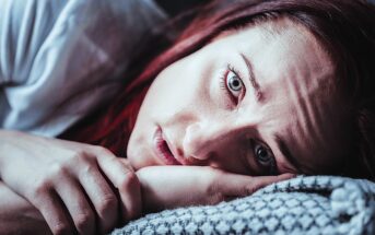 A woman with red hair lies on her side, resting her head on a patterned pillow. Her expression is solemn, and her hand is near her face. The image has a blueish tint, emphasizing a moody atmosphere.