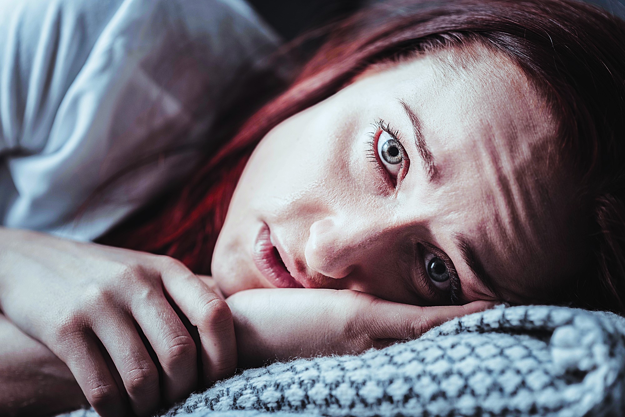 A woman with red hair lies on her side, resting her head on a patterned pillow. Her expression is solemn, and her hand is near her face. The image has a blueish tint, emphasizing a moody atmosphere.