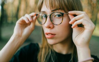 A person with long hair and glasses is adjusting their frames while standing outdoors. The background is a blurred forest, creating a natural, serene setting. The person appears thoughtful and focused.