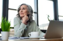 An elderly woman with gray hair sits at a desk, gazing thoughtfully out of the window. She rests her chin on her hand, and a laptop, cup, and small plant are on the desk. Sunlight streams in through large windows in the background.