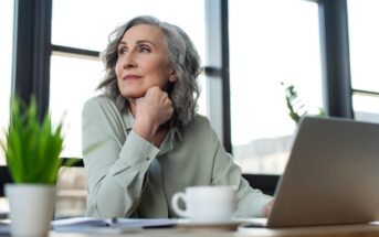 An elderly woman with gray hair sits at a desk, gazing thoughtfully out of the window. She rests her chin on her hand, and a laptop, cup, and small plant are on the desk. Sunlight streams in through large windows in the background.