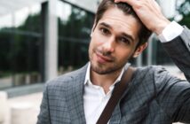 A man in a gray plaid suit and white shirt stands outdoors, touching his hair with one hand. He has a slight smile and is carrying a shoulder bag. The background features a blurred glass building and trees.