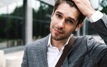 A man in a gray plaid suit and white shirt stands outdoors, touching his hair with one hand. He has a slight smile and is carrying a shoulder bag. The background features a blurred glass building and trees.