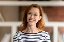 A person with red hair is smiling and wearing a black and white striped shirt. They're in a room with blurred bookshelves in the background.