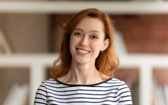 A person with red hair is smiling and wearing a black and white striped shirt. They're in a room with blurred bookshelves in the background.