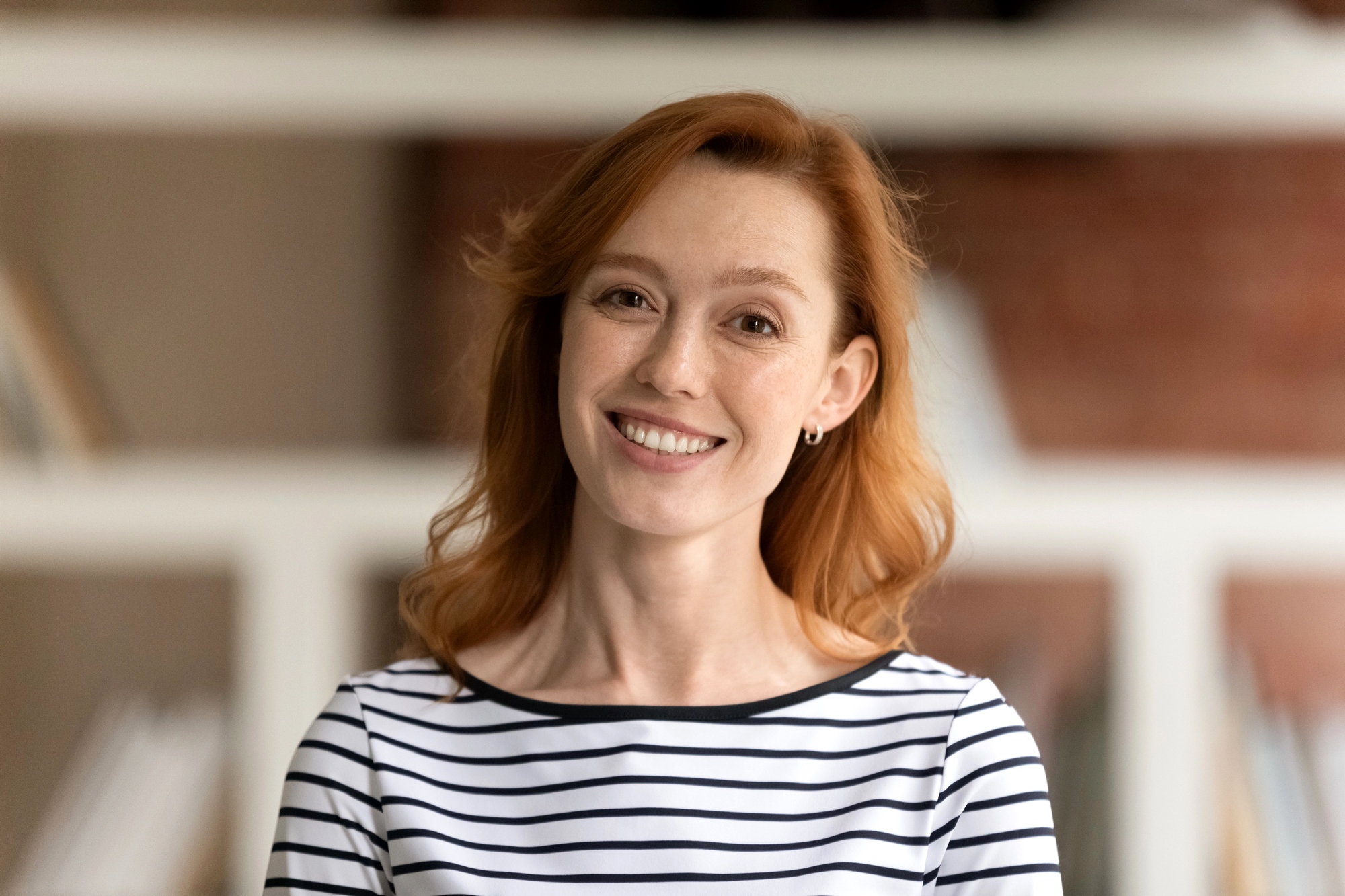 A person with red hair is smiling and wearing a black and white striped shirt. They're in a room with blurred bookshelves in the background.