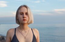 A person with short blond hair is standing on a beach, wearing a dark swimsuit and a necklace. The background shows a calm sea and a cloudy sky.