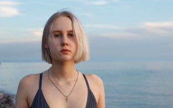 A person with short blond hair is standing on a beach, wearing a dark swimsuit and a necklace. The background shows a calm sea and a cloudy sky.