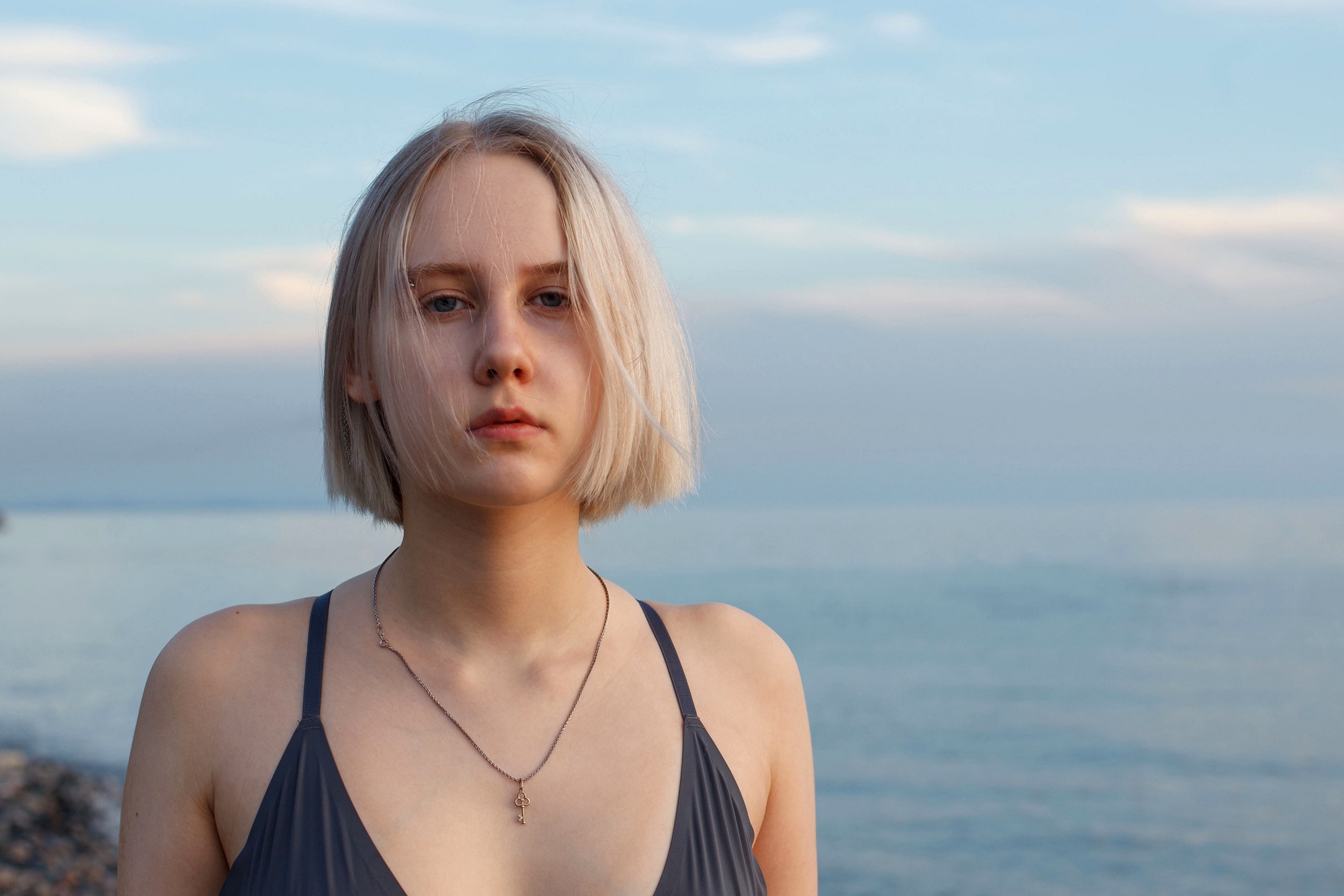 A person with short blond hair is standing on a beach, wearing a dark swimsuit and a necklace. The background shows a calm sea and a cloudy sky.