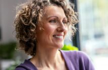 Smiling woman with curly hair wearing a purple top, looking out a window in a bright room.