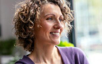 Smiling woman with curly hair wearing a purple top, looking out a window in a bright room.