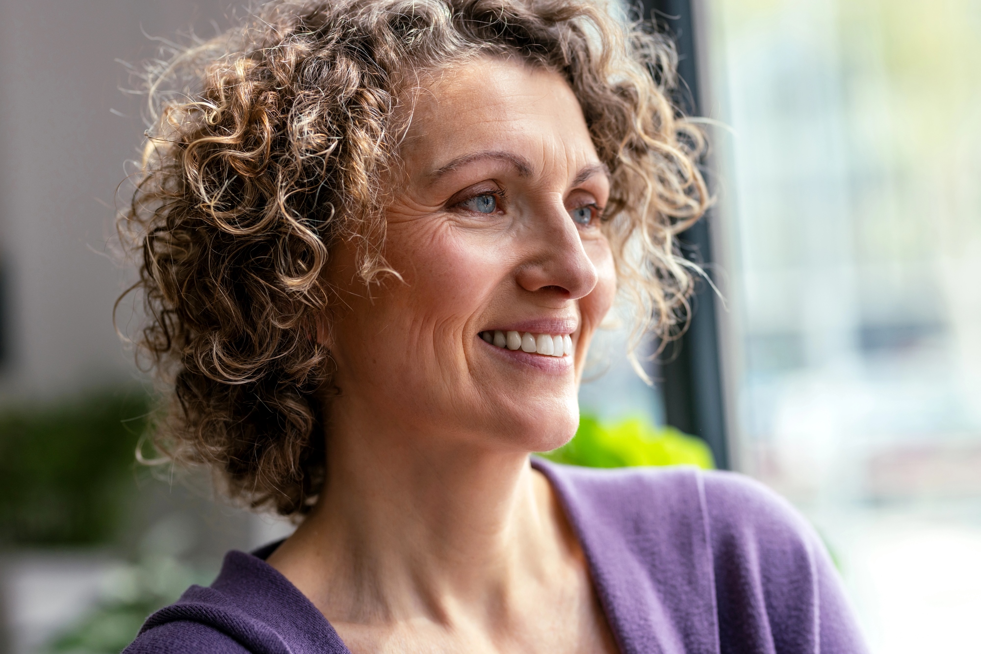 Smiling woman with curly hair wearing a purple top, looking out a window in a bright room.