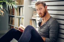 A man with a beard and long hair tied back is sitting on the floor, leaning against a bookshelf. He's wearing a gray long-sleeve shirt and dark pants, holding an open book, with a thoughtful expression.