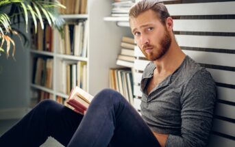 A man with a beard and long hair tied back is sitting on the floor, leaning against a bookshelf. He's wearing a gray long-sleeve shirt and dark pants, holding an open book, with a thoughtful expression.