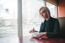 A person with blonde hair sits at a table in a cafe, holding a white cup and saucer. They are wearing a black and white polka dot shirt with a black vest. A large window shows an urban cityscape outside.