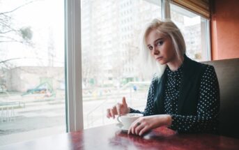 A person with blonde hair sits at a table in a cafe, holding a white cup and saucer. They are wearing a black and white polka dot shirt with a black vest. A large window shows an urban cityscape outside.