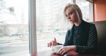 A person with blonde hair sits at a table in a cafe, holding a white cup and saucer. They are wearing a black and white polka dot shirt with a black vest. A large window shows an urban cityscape outside.