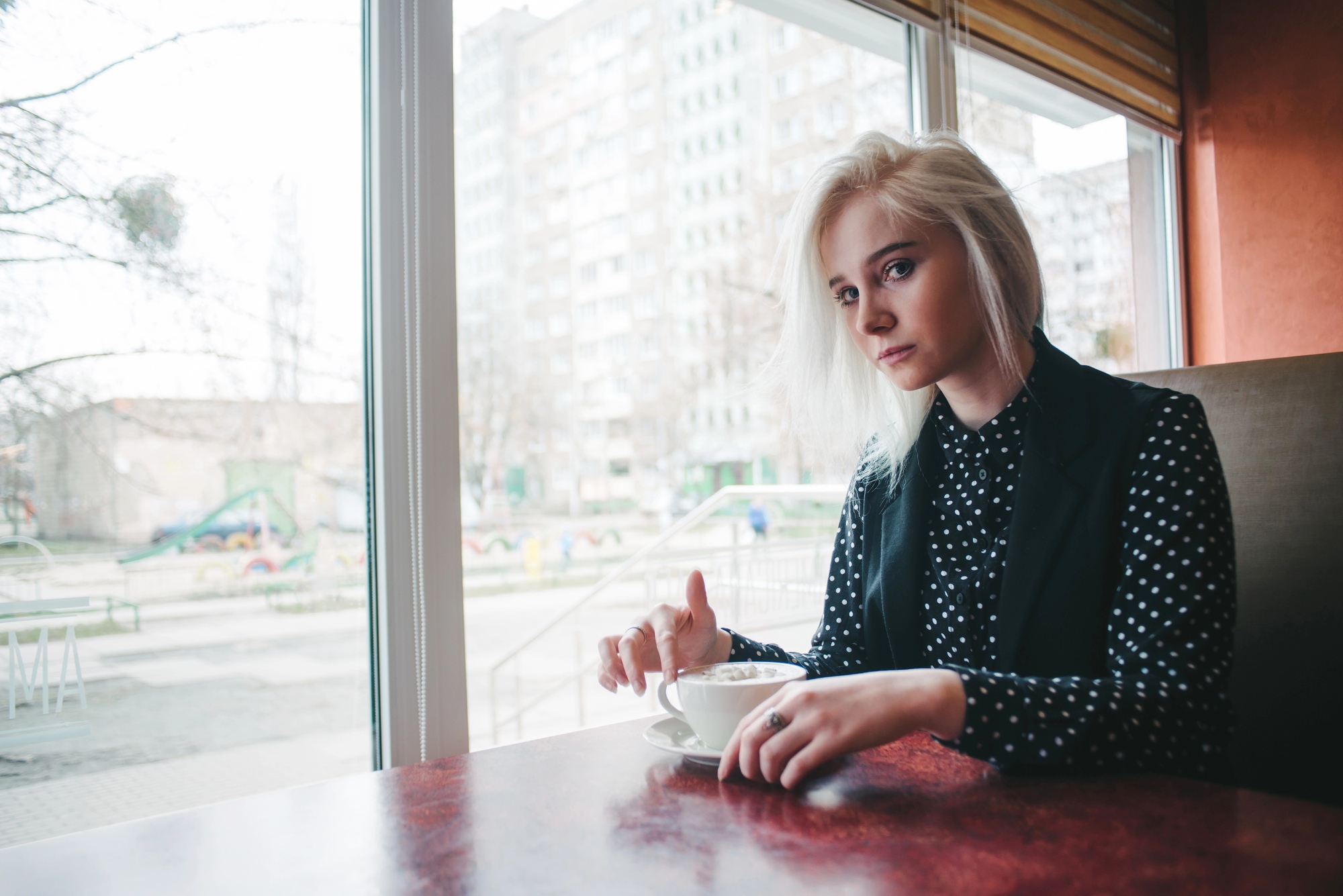 A person with blonde hair sits at a table in a cafe, holding a white cup and saucer. They are wearing a black and white polka dot shirt with a black vest. A large window shows an urban cityscape outside.