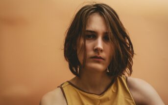 A person with shoulder-length brown hair is wearing a sleeveless yellow top, looking towards the camera with a serious expression. The background is a gradient of warm orange tones.