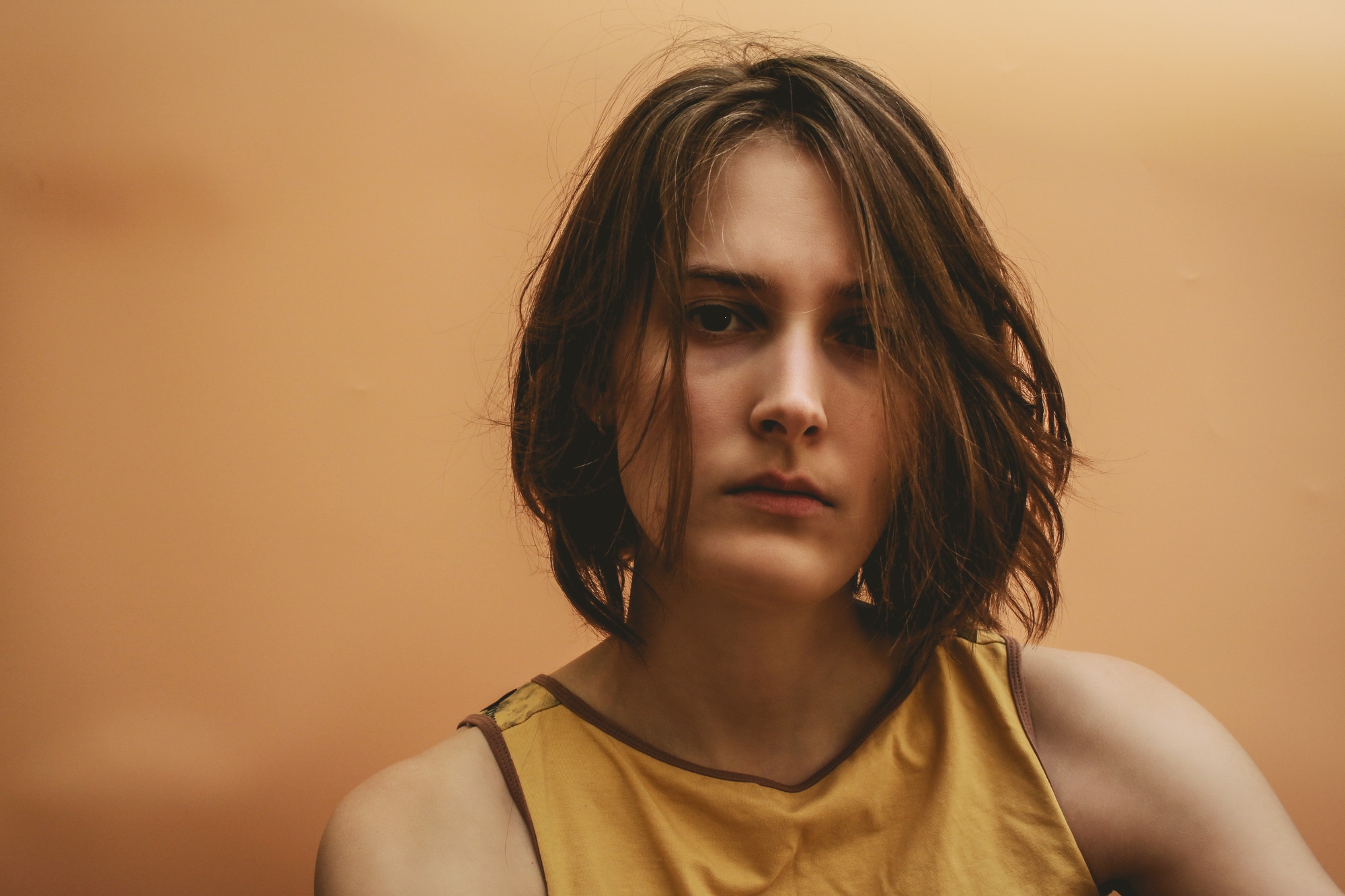 A person with shoulder-length brown hair is wearing a sleeveless yellow top, looking towards the camera with a serious expression. The background is a gradient of warm orange tones.