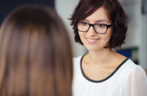 A woman with shoulder-length brown hair and glasses is smiling while talking to another person who is facing away from the camera. She is wearing a white blouse with black trim, and the background appears to be a blurred indoor setting.