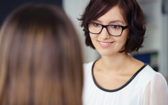 A woman with shoulder-length brown hair and glasses is smiling while talking to another person who is facing away from the camera. She is wearing a white blouse with black trim, and the background appears to be a blurred indoor setting.